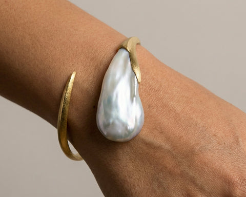 Gold bracelet with a large pearl on a wrist against a neutral background