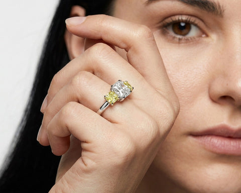 Close-up of a woman's hand wearing a ring with a yellow gemstone and diamond, against a neutral background.