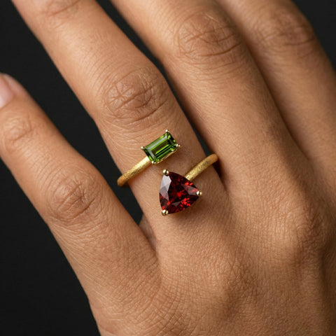 Two gold rings with green and red gemstones on a hand against a dark background