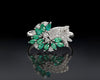 Elegant ring with emerald green gemstones and silver accents on a reflective surface.