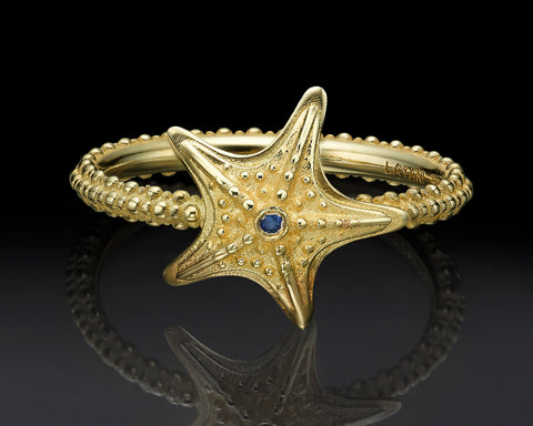 Starfish ring in 14K yellow gold with a blue sapphire, lying flat to show the starfish from above, textured with raised gold granules