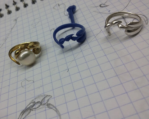 Finished gold ring beside its blue wax carving, silver model and original sketch